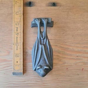 May include: A cast metal bat-shaped door knocker with a detailed design. The knocker is a dark grey colour and is approximately 15 cm long. The bat is hanging upside down, with its wings folded. The knocker is mounted on a wooden surface.