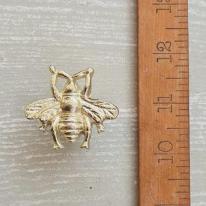May include: A gold-toned bee-shaped metal object positioned next to a wooden ruler. The bee features detailed wings and a segmented body. The ruler shows measurements in centimetres, indicating the bee is approximately 3.8 centimetres in width.