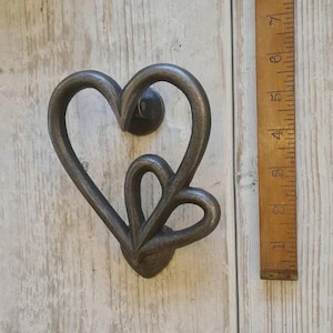 May include: A dark grey metal heart-shaped door knocker. The knocker is approximately 15 cm tall and features a double heart design. It is mounted on a weathered, light-coloured wooden surface.