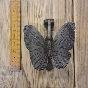 May include: A dark grey cast iron butterfly door knocker with detailed wing patterns. The door knocker is mounted on a wooden door. A wooden ruler is next to the door knocker, measuring approximately 13 cm.