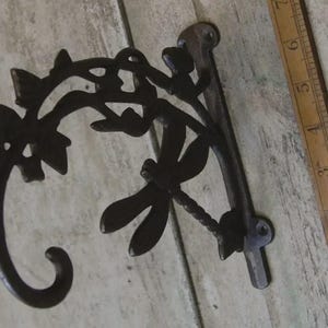 May include: A dark brown, cast iron wall-mounted plant hanger. The decorative bracket features a dragonfly and leaf design. A wooden ruler is next to the hanger. The hanger is designed to hold a hanging basket or other decorative items.