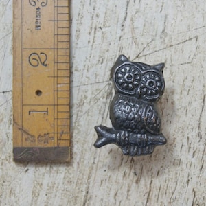 May include: A dark grey metal owl figurine perched on a branch, positioned next to a wooden ruler showing a height of approximately 5 cm. The owl features textured feather details and large circular eyes with a distinct, raised relief pattern.
