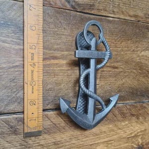 May include: A dark grey metal anchor with a rope design, approximately 15 cm tall, sits on a wooden surface. The anchor has a ring at the top and a detailed design. A wooden ruler is positioned to the left of the anchor.