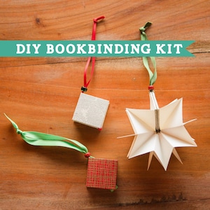 May include: A DIY BOOKBINDING KIT featuring three hanging ornaments: a small silver square, a small red plaid square, and a white folded paper star. Each ornament includes a colored ribbon loop for hanging, with red or green fabric ribbons.