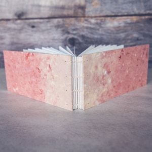 May include: A closed book with a pink and white marbled cover. The book is bound with a white thread and has a spine with a decorative stitch.