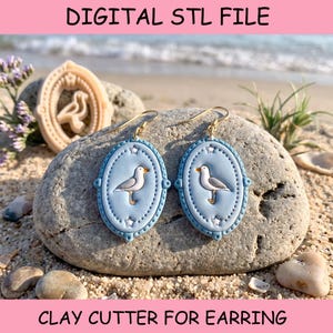 May include: A pair of oval-shaped earrings in light blue with a white and grey seagull graphic, featuring gold-tone hooks. The earrings have a beaded border with small star accents. The text DIGITAL STL FILE CLAY CUTTER FOR EARRING is visible.