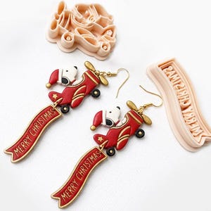 May include: A pair of dangle earrings featuring a white dog in a red Santa hat piloting a red airplane, attached to a red banner with the text Merry Christmas in gold. Two light-colored plastic cookie cutters are included, one shaped like the airplane.