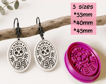 May include: A digital STL file for a clay cutter for earrings, featuring a skull design. The set includes three sizes: 35mm, 40mm, and 45mm. Two oval-shaped white earrings with black skull illustrations and black metal hooks are displayed alongside a purple cutter.
