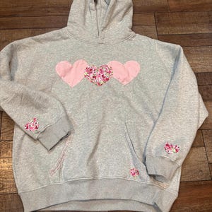 May include: A gray hooded sweatshirt with three pink hearts sewn on the front. The hearts have a floral pattern. There are also smaller floral hearts sewn on the sleeves and the pocket.