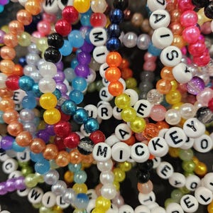 May include: A collection of colourful beaded bracelets featuring white letter beads spelling out names like Mickey, Daisy, and Cinderella. The bracelets are made of various round, glossy, and translucent plastic beads in shades of blue, pink, yellow, and orange.