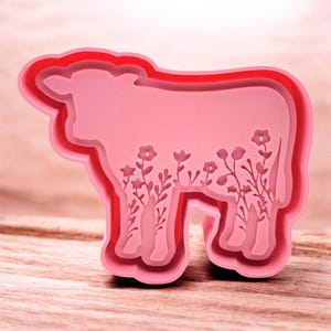 May include: A pink cow-shaped cookie cutter with a floral design. The cutter has a layered design with a red outline and detailed floral cutouts within the cow's body. The cutter is made of plastic and is designed for baking.