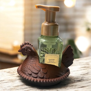 May include: A brown chicken-shaped soap holder cradles a green soap bottle with a pump. The bottle has a label that reads "Waikiki Beach Coconut". The holder is made of a dark brown material and sits on a wooden surface.
