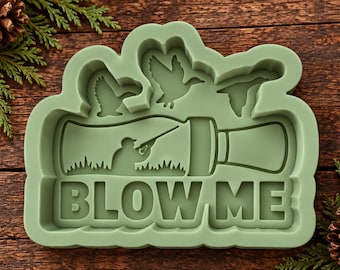May include: A sage green silicone mold featuring a raised design of a duck call, a hunter aiming a shotgun, and three flying ducks. The text BLOW ME is embossed in large capital letters at the bottom of the mold, which has a wavy outer edge.