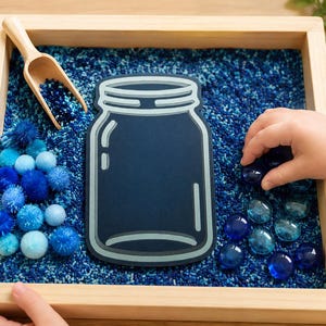 May include: A wooden tray filled with blue sensory materials, including pom-poms, glass beads, and a wooden scoop. A large, dark blue jar-shaped cutout is in the centre. A child's hand is reaching for the glass beads.