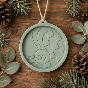May include: A sage green circular ornament featuring a raised relief design of a central circle with two small inner rings, surrounded by curved lines and a grid of squares. The ornament hangs from a thin, light brown jute twine string.