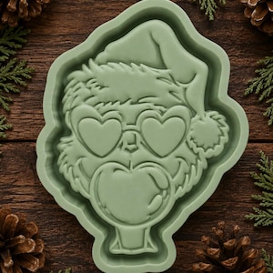 May include: A sage green silicone mold featuring a detailed embossed design of a furry creature wearing a Santa hat, heart-shaped glasses, and blowing a large bubble. The mold has a smooth, matte finish and a raised border around the central figure.