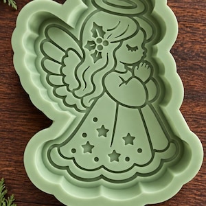 May include: A sage green silicone mold featuring a recessed design of an angel with a halo, wings, and a dress decorated with stars. The angel is depicted with hands clasped in prayer and a holly sprig detail in the hair, creating a clear impression.