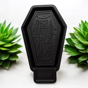 May include: A black, coffin-shaped decorative object with a textured surface. The center features an embossed bat icon and the text "JUST A LITTLE RAY OF PITCH BLACK" in capital letters, surrounded by small star and diamond-shaped accents.