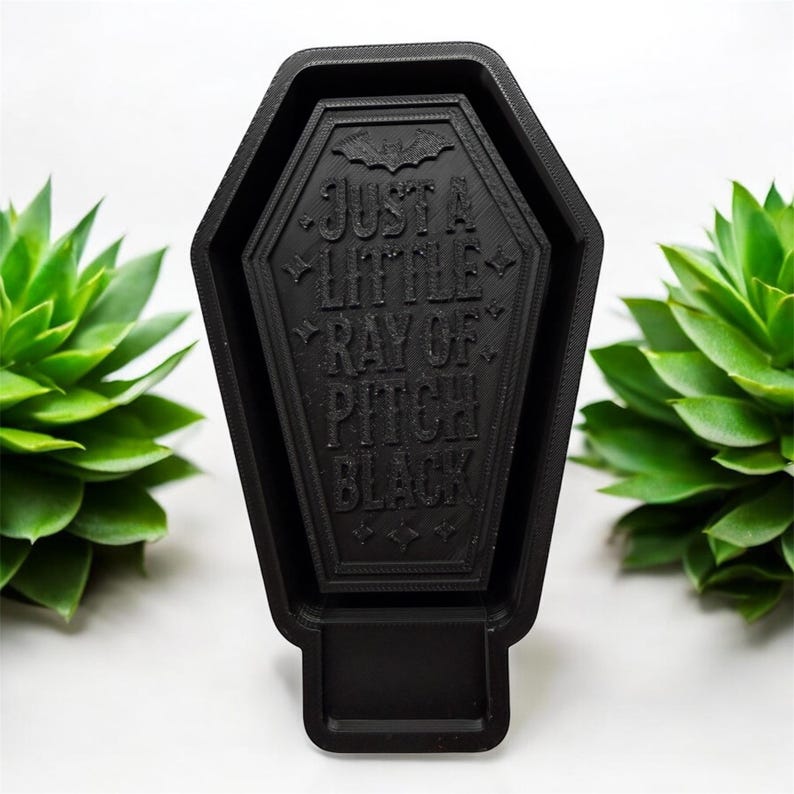 May include: A black, coffin-shaped decorative object with a textured surface. The center features an embossed bat icon and the text "JUST A LITTLE RAY OF PITCH BLACK" in capital letters, surrounded by small star and diamond-shaped accents.