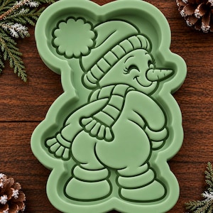 May include: A light green silicone mold featuring a raised, embossed design of a snowman wearing a pom-pom hat and a scarf. The snowman is depicted in profile, looking over its shoulder with a carrot nose, while wearing boots and mittens.