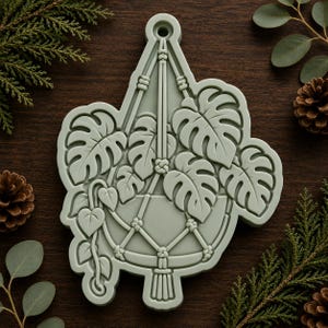 May include: A pale sage green silicone mold featuring a detailed embossed design of a hanging plant in a macrame planter. The design includes multiple monstera leaves and trailing vines, with a circular hanging loop at the top center.