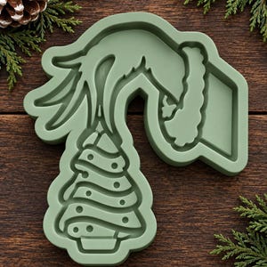 May include: A sage green silicone mold featuring a recessed design of a stylized hand reaching down to place a star atop a Christmas tree, with a Santa hat silhouette integrated into the upper right section of the mold's frame.