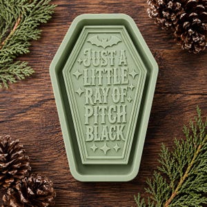 May include: A sage green silicone mold shaped like a coffin with a raised bat icon at the top and the text "JUST A LITTLE RAY OF PITCH BLACK" embossed in the center, surrounded by small four-pointed star accents throughout the design.