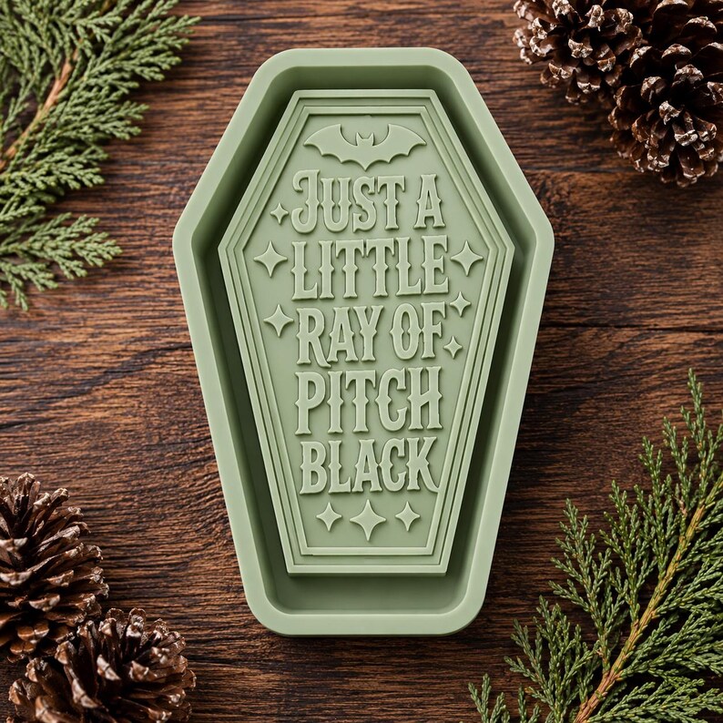 May include: A sage green silicone mold shaped like a coffin with a raised bat icon at the top and the text "JUST A LITTLE RAY OF PITCH BLACK" embossed in the center, surrounded by small four-pointed star accents throughout the design.
