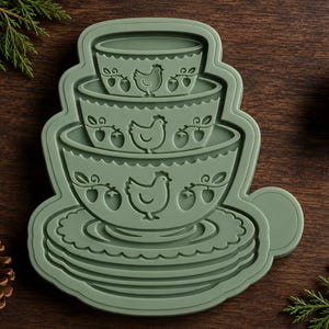 May include: A sage green cookie cutter featuring a stacked design of three bowls and a plate. Each bowl is embossed with a chicken and strawberry motif, while the base plate has a scalloped edge. The cutter has a smooth, molded plastic finish.