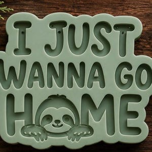 May include: A sage green silicone mold featuring the embossed text I JUST WANNA GO HOME with a small sloth character integrated into the letter O. The mold has a smooth, matte finish and a rounded, cloud-like border shape for crafting projects.
