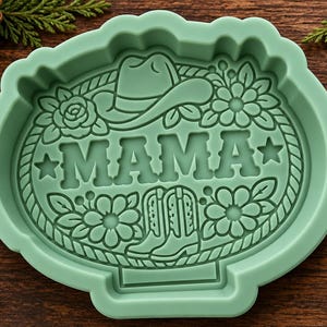 May include: A light green silicone mold featuring a central embossed design of the word MAMA, a cowboy hat, a pair of cowboy boots, and floral accents, all enclosed within a rope-style border. The mold has a scalloped edge and a flat base.