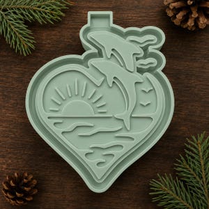 May include: A sage green heart-shaped cookie cutter featuring an embossed design of two dolphins leaping above ocean waves with a sun rising on the horizon and two small birds flying in the sky, all contained within a heart-shaped outline.