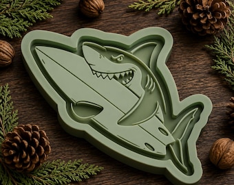May include: A sage green silicone mould featuring a raised, detailed design of a shark biting into a surfboard. The mould has a smooth, matte finish and a contoured edge that follows the shape of the shark and board illustration.