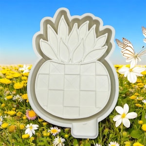 May include: A white pineapple-shaped cookie cutter with a textured surface. The cutter features a detailed design of a pineapple, including the crown and the checkered body. The background shows a field of yellow flowers and butterflies.