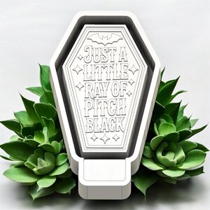 May include: A white coffin-shaped object with a flat top featuring the engraved text 'JUST A LITTLE RAY OF PITCH BLACK' and a small bat icon at the top. The design includes decorative star accents and clean, geometric lines throughout the surface.