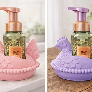 May include: Two chicken-shaped soap holders, one pink and one purple, each holding a soap dispenser. The dispensers have bronze pumps and labels reading "Waikiki Beach Coconut". The holders are ceramic.
