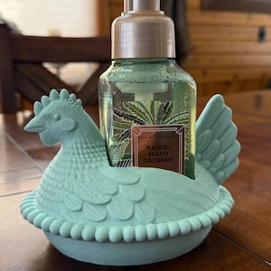 May include: A light green chicken-shaped soap dispenser holder with a bottle of soap inside. The soap bottle has a label that reads "Waikiki Beach Coconut". The holder is made of a solid material and sits on a wooden surface.