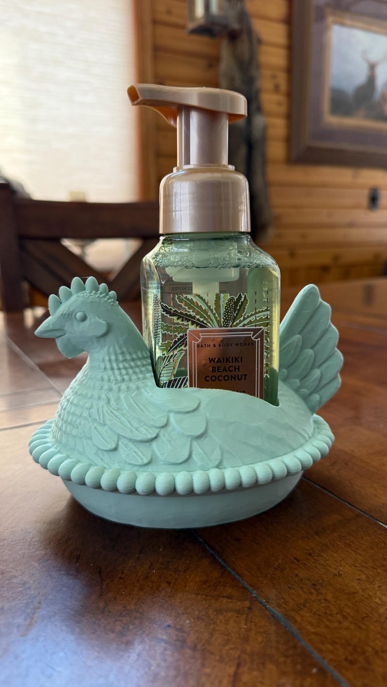 May include: A light green chicken-shaped soap dispenser holder with a bottle of soap inside. The soap bottle has a label that reads "Waikiki Beach Coconut". The holder is made of a solid material and sits on a wooden surface.