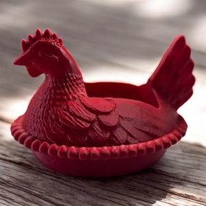 May include: A red, hen-shaped decorative container. The container is designed to resemble a hen sitting on a nest. The hen has detailed feathers, a comb, and a beak. The container is made of a solid material and is suitable for holding small items.