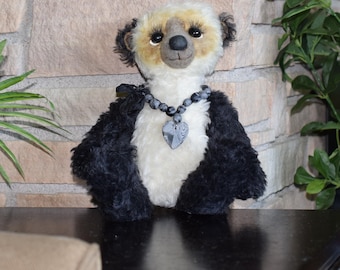 May include: A handmade panda bear plush toy with a cream-colored face and chest, black fur on the arms and ears, and a black bead necklace with a heart-shaped pendant. The bear is posed on a dark surface.