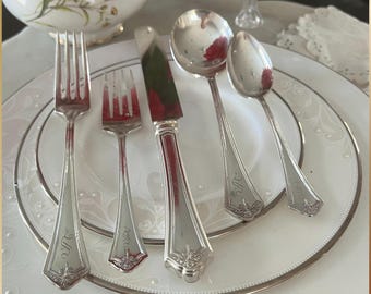 May include: Silverware set on a white plate with a silver rim. The set includes a fork, a smaller fork, a knife, a spoon, and a smaller spoon. Each piece has a decorative handle and an engraved initial.
