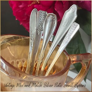 Può includere: Una collezione di cucchiaini vintage argentati con disegni ornati, esposti in una brocca di vetro color oro. I cucchiaini presentano vari motivi floreali e geometrici. Il testo in basso recita "Vintage Mix and Match Silver Plate Small Spoons."