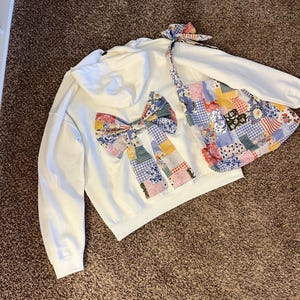 May include: White hoodie with a large patchwork bow on the back and a matching bow on the strap of a patchwork bag. The patchwork features floral, gingham, and solid patterns in various colours.