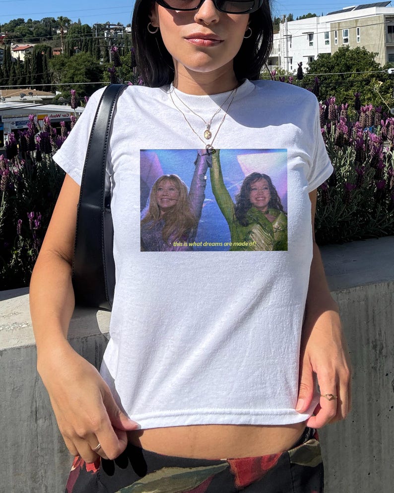 May include: White t-shirt with a rectangular image of two women with raised arms. The image has a purple and blue background and the text "this is what dreams are made of." The shirt is a casual style.