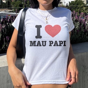 May include: White t-shirt with the text "I ❤️ MAU PAPI" in black and red. The shirt has a crew neck and short sleeves. The image shows the shirt being worn outdoors on a sunny day.
