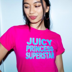 May include: A bright pink t-shirt with the words "JUICY PRINCESS SUPERSTAR" in turquoise block letters. The shirt has short sleeves and a crew neck. The model is wearing hoop earrings.