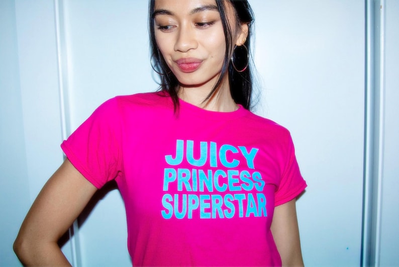 May include: A bright pink t-shirt with the words "JUICY PRINCESS SUPERSTAR" in turquoise block letters. The shirt has short sleeves and a crew neck. The model is wearing hoop earrings.