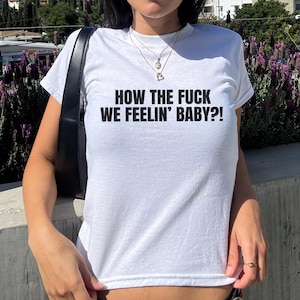 May include: White t-shirt with the text "HOW THE FUCK WE FEELIN' BABY?!" in black. The shirt is short-sleeved and appears to be made of a soft material. The person is wearing sunglasses and necklaces.