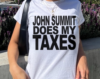 May include: A white short-sleeved t-shirt featuring the bold black text "JOHN SUMMIT DOES MY TAXES" printed across the chest. The garment has a crew neckline and a standard fit, showcasing clear, high-contrast lettering on a plain white fabric base.