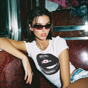 May include: A white t-shirt featuring a black and white lip graphic with a pink heart. The person is wearing dark sunglasses and a silver chain necklace. The background shows a red and brown interior.